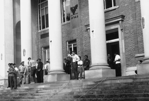 Jesus Salas, in a white shirt and standing in front of the microphone with his arm raised, addresses a rally at the top of the steps in front of Waushara Courthouse. Above him hangs a National Farm Workers Association (NFWA) banner with an Aztec eagle symbol at the center. Salas is the leader of Oberos Unidos (United Workers,an independent farm worker labor union effort in the 1960s. Salas is between two columns with Obreros Unidos signs that say "Juntarnos para ser reconocidos. Hablar para ser oidos. La raza tiene causa"/ "Join to be recognized. Speak to be heard. The people have a cause." The column on the left has an image of our Lady of Guadalupe. Behind and to the left of Salas are Obreros Unidos members and supporters wearing Obreros Unidos pins with the eagle symbol.<p>Salas expresses the demands of migrant farm workers to provide them with $1.25 minimum wage, insurance benefits, a meeting with the governor's committee, and public sanitation facilities in Wautoma.<p>Strikes, rallies and marches in Wisconsin were organized by Obreros Unidos, an independent farm worker labor union effort in the 1960s<p>Jesús Salas en la manifestación enfrente del tribunal de Waushara<p>Jesús Salas, vestido en una camisa blanca y parado enfrente de un micrófono con su brazo alzado, se dirige al público enfrente del tribunal o Waushara Courthouse. Arriba de él cuelga la bandera de la Asociación Nacional de Trabajadores Agrícolas (NFWA, por sus siglas en inglés) con el símbolo de una águila Azteca en el centro. Salas es el líder de Obreros Unidos (un sindicato independiente de trabajadores agrícolas durante los años 1960). Salas está en medio de dos columnas con letreros de Obreros Unidos que dicen, "Juntarnos para ser reconocidos. Hablar para ser oídos. La raza tiene causa." La columna a la izquierda tiene una imagen de la Virgen de Guadalupe. Detrás y hacia la izquierda de Salas, están miembros de Obreros Unidos y simpatizantes con botones de apoyo con el símbolo de una águila. Salas expresa las demandas de los trabajadores agrícolas emigrantes, que piden un sueldo mínimo de $1.25 por hora, beneficios de aseguranza, una junta con el comité del gobernador, e instalaciones sanitarias públicas para los trabajadores en Wautoma. Las huelgas, manifestaciones, y marchas en Wisconsin fueron organizadas por Obreros Unidos, un esfuerzo independiente del sindicato de trabajadores agrícolas durante los años 1960.</p>