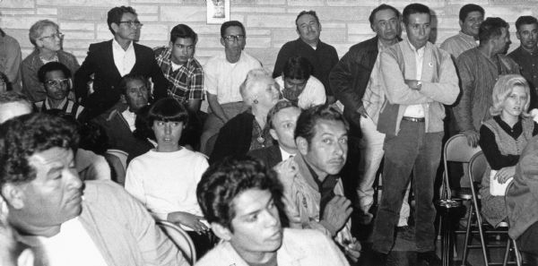 Obreros Unidos (United Workers) leaders, members and supporters at an Industrial Commission Hearing at Waushara County Courthouse. On the right, standing with his arms folded and wearing a jacket is Salvador Sanchez, union leader. To his left, seated with his head down is Jesus Salas, also a union leader. The Waushara County Courthouse hearing discussed the issue of paying minimum wages to migrant farm workers.<p>Strikes, rallies and marches in Wisconsin were organized by the Obreros Unidos, an independent farm worker labor union effort in the 1960s.<p>Obreros Unidos en la audiencia pública de la Comisión Industrial<p>Lideres, miembros y simpatizantes de Obreros Unidos en la audiencia pública de la Comisión Industrial en el tribunal del condado o Waushara County Courthouse. A la derecha, parado con sus brazos cruzados y vistiendo una chaqueta está Salvador Sánchez, líder de sindicato. A su izquierda, sentado con su cabeza baja está Jesús Salas, también un líder de sindicato. En la audiencia pública en el tribunal del condado o Waushara County Courthouse se discutió la cuestión de pagarles un salario mínimo a los trabajadores agrícolas emigrantes. Las huelgas, manifestaciones, y marchas en Wisconsin fueron organizadas por Obreros Unidos, un esfuerzo independiente del sindicato de trabajadores agrícolas durante los años 1960.</p>