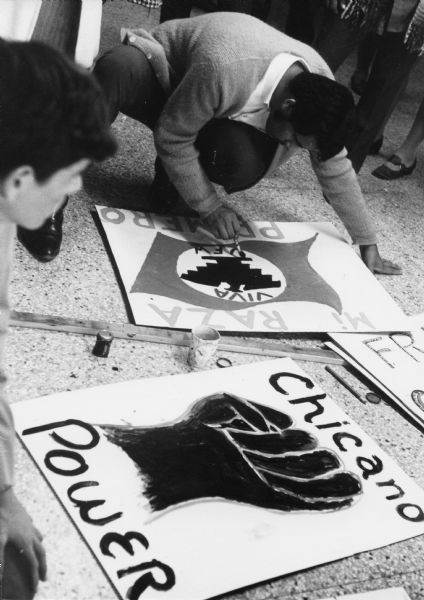 Obreros Unidos (United Workers) demonstrators painting signs at a Milwaukee rally. In the background, a man is painting the National Farmworker's (NFW) Aztec eagle symbol on a poster. The message on the  poster says: "Mi raza primero/Viva NFW (My race first/Long live NFW)." Another sign on the ground says "Chicano Power" with a raised fist symbol at its center.<p>This photograph is a part of Wisconsin-native David Giffey's series "Struggle for Justice," images from the migrant farm worker struggle including an independent organizing effort in Wisconsin and the nationwide grape boycott movement started by Cesar Chavez of United Farm Workers during the 1960s and 1970s.<p>Letreros para una demostración<p>Demostradores de Obreros Unidos están pintando unos letreros durante una manifestación en Milwaukee. Hacia el fondo, un hombre está pintando en un póster el símbolo usado por National Farmworkers (NFW) de una águila Azteca. El mensaje en el póster dice: "Mi raza primero/Viva NFW." Otro letrero en el suelo dice "Poder Chicano" ("Chicano Power") con el símbolo de un puño al centro. Esta fotografía es parte de la serie "Lucha por la Justicia" tomada por David Giffey, originario de Wisconsin, las imágenes muestran la lucha de los trabajadores agrícolas emigrantes incluyendo un esfuerzo independiente organizado en Wisconsin y el movimiento nacional del boicot de uvas empezado por Cesar Chávez de la unión de campesinos o United Farm Workers durante los años 1960 y 1970.</p>