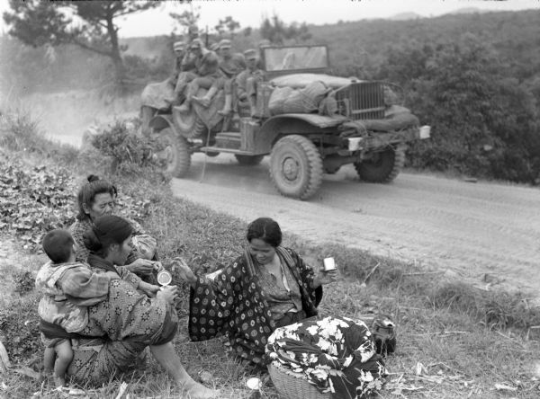 A military truck with men in uniform is on a road near three Japanese women and a small child who are sitting on the grass on the roadside. Two of the  women are holding small metal containers, and a basket with a flowered cloth in it is beside them. The image was taken in Okinawa, Japan.