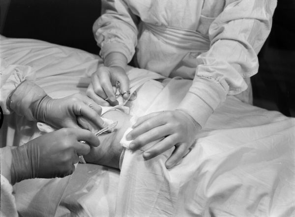 View of the gloved hands of a pair of surgeons as they begin to operate on a patient in Japan. One person is holding a scalpel over a portion of an exposed ankle, the other is holding a pair of scissors. Both surgeons are also holding gauze on the exposed skin. The rest of the body is covered by white cloth. Made on the hospital ship USS <i>Relief</i> trip for a typhoon.