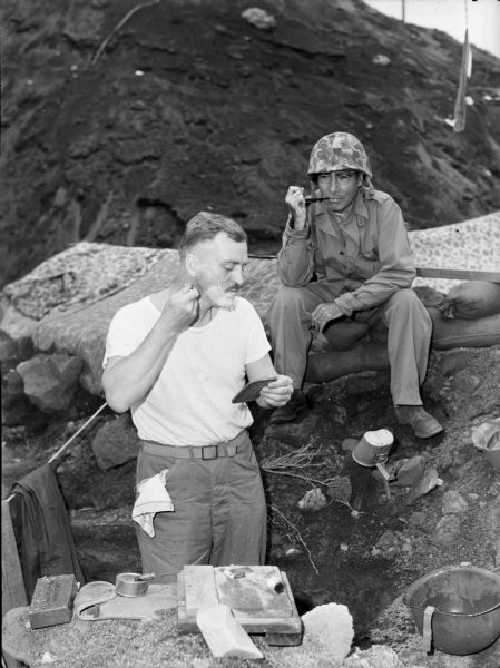 Commander A.E. Reymont shaves his face while division surgeon Captain C.P. Archambault looks on. Captain C.P. Archambault, Medical Corps, United States Navy Reserve, III Division surgeon, sits on sandbags holding a pipe in his mouth. He wears fatigues, a camouflage helmet, and glasses. Commander Reymont wears a white t-shirt and pants with a cloth hanging out of the pocket. Behind him is a clothesline with clothing on it. He holds a mirror in one hand and a shaving razor in the other. He has shaving cream on half of his face. The foxhole became quarters for doctors and commanding officers.