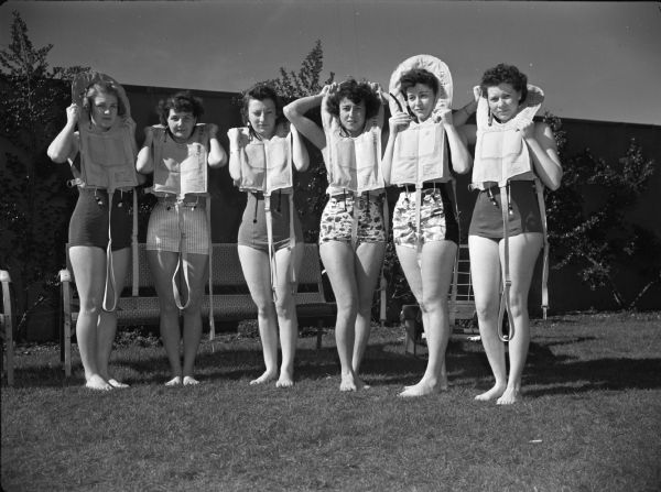 Six female flight nurses posing outdoors. The women are wearing bathing suits and holding life vests around their necks at the at Alameda Naval Air Strip during Aquatic Training.