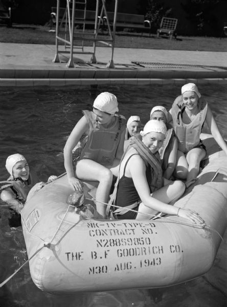 Six female flight nurses in a swimming pool during aquatic training at the at Alameda Naval Air Strip. Four of the women are in a raft, and two women are in the water. All the women are wearing bathing suits, bathing caps, and life vests.