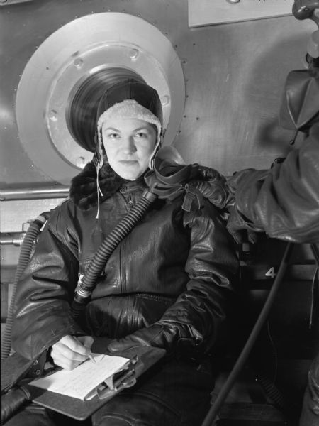 Gwen Jensen, a female flight nurse, poses for a photograph during flight training at Alameda Naval Air Strip. In an anoxic faint chamber, used to simulate high altitude flying with little oxygen, Gwen Jensen writes on a clipboard as another person holds a mask towards her face. She is wearing heavy leather clothing, including a hat and glove on one hand. Her leather jacket has a black fur collar. Tubing for the mask crosses her chest.