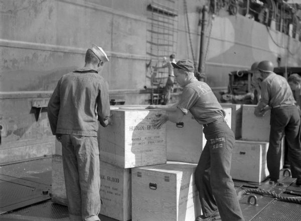 Two men working together to move a box marked "Human Blood" at Buckner Bay, Okinawa, Japan. They are aboard a tank landing ship pontoon float. One man is wearing a shirt with the sleeves rolled up, pants with the name "Oliver" written on them, and a brimmed cap. The other man is wearing a uniform and a sailor hat. Another man in the background is wearing a helmet. The side of the ship is in the background.