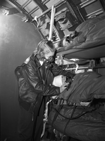 A female flight nurse during flight training with patients. She is leaning over the patient in a bunk bed to place a mask. The student is wearing a heavy leather jacket and pants. She is also wearing a mask over part of her face with a long tube attached to the bottom. Beside her are bunk beds with two people in sleeping bags. The person she is putting the mask on has a head wrapped in bandages. The side of the inside of the plane is in the background. The photograph was taken in flight at Alameda Navy Air Strip.