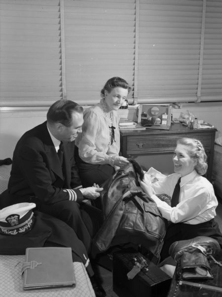 Flight nurse, Gwen Jensen, visiting with her parents as she prepares to leave U.S. soil. Jensen is kneeling on the floor as her parents are sitting on a bed next to her. A heavy leather jacket is laid across the collection of bags on the floor. Jensen and her parents are all holding on to part of the jacket. A military hat is on the bed on top of a military jacket. Jensen is smiling, as is her father. A toiletry case is on the bedside table. Jensen is wearing a collared shirt and tie. Her father is in a military uniform. Jensen's mother is wearing a striped shirt with a ruffled collar.