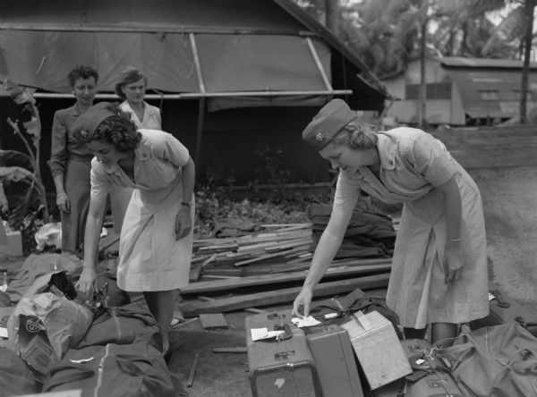 Flight nurses, newly arrived in Agana Airport, Guam, searching for their luggage as two other women are looking on. The two flight nurses have just completed flight training in Alameda, California. Both women are wearing light dresses and caps. Cabins and palm tress are in the background.