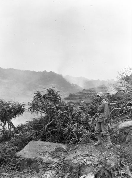 The front at Nago, Okinawa.  A uniformed soldier stands on a rocky outcropping near bushes.  Misty or smoky hillsides are visible in the background.