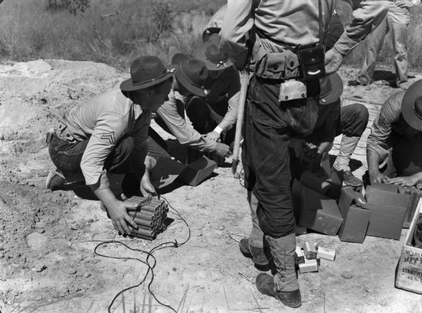 Trainees at Rio Hato Infantry Artillery School setting dynamite charges outdoors in Panama. The men are wearing brimmed hats and uniforms. One crouching man is holding onto a bundle of dynamite sticks. Other men are looking through boxes.