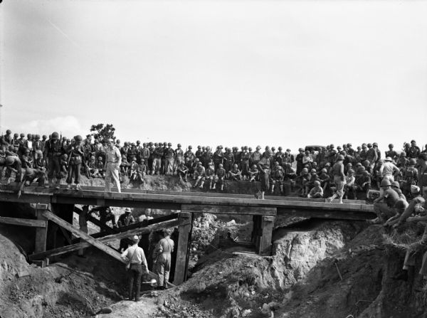 Trainees at Rio Hato Infantry Artillery School, Panama, gathered around a wooden bridge they are rigging with dynamite charges. The men are wearing fatigues and some are carrying sub-machine guns. One man in the foreground is holding a camera near other men underneath the bridge.