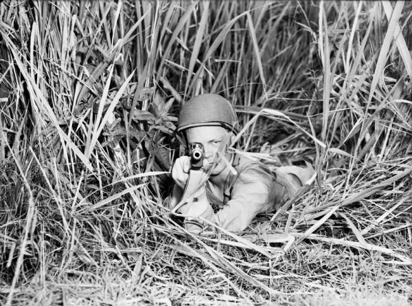 A soldier from the 14th Infantry points a submachine gun at the camera. He is laying on his stomach with tall grass around him. He is wearing fatigues and a helmet. This photograph was taken in Panama.