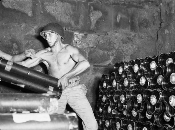 A soldier stacks ammunition for a coastal gun in Panama near a stone wall. The soldier is being handed a case of ammunition. He is wearing a helmet and pants, but no shirt.