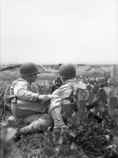 View from behind towards two soldiers lying on a hill among shrubs overlooking a hilly landscape with a submachine gun. The soldiers are wearing fatigues and helmets.