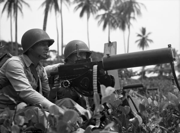 A two-man crew mans a .30mm machine gun in Panama. The men are wearing fatigues. There are trees in the background and plants in the foreground.