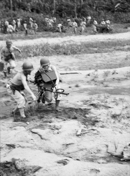 A machine gun crew running with their .30 machine gun. Two men in the foreground are wearing pants and helmets but no shirts, and are carrying a machine gun between them. Another man is running behind them. In the background a group of men near scrub are looking towards them. This photograph was taken in Panama.