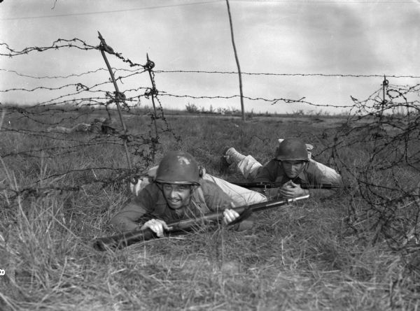 Two men posing during a training exercise on cutting barbed wire at Rio Hato Infantry Artillery School, Panama. The men are on their stomachs crawling through grass next to two fences made of barbed wire. The men are wearing fatigues and carrying submachine guns. Another man can be seen in the background also crawling on his stomach.