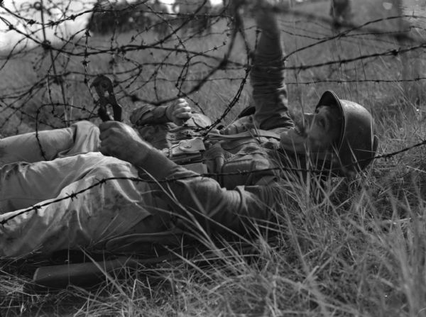 Two male soldiers wearing fatigues cutting barbed wire during a training exercise at Rio Hato Infantry Artillery School in Panama. The men are on their backs on the grass with barbed wire above and around them. One man is holding a wire cutter and a rifle is on the ground next to him.