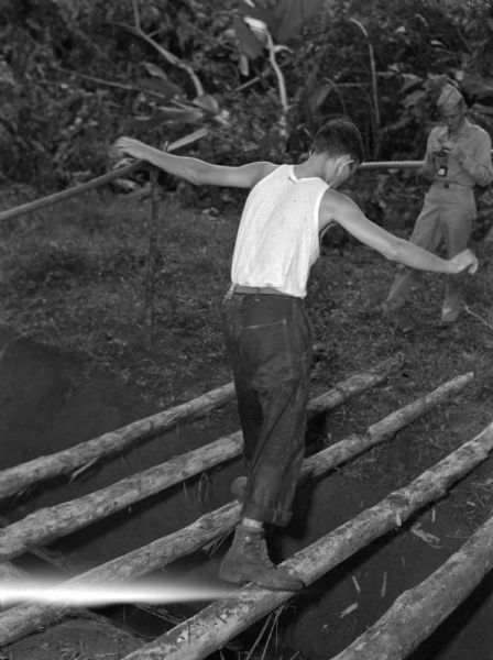 A soldier from the 14th Infantry balancing on an obstacle course at Fort Davis, in Panama. The soldier is balancing on a wooden log, and is wearing a sleeveless white shirt, rolled up pants, and boots. He has his arms out for balance as he is crossing to another log. Another man in uniform is on the other side of the ditch.