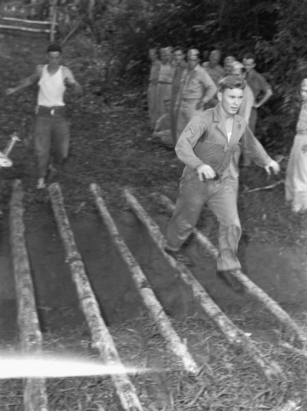 Two men from the 14th Infantry are running over logs on an obstacle course at Fort Davis, in Panama. One man is wearing a jumpsuit and the other man is wearing a sleeveless t-shirt and pants. The logs they are running over are set across a dirt ditch. A group of men in uniforms watch them near trees in the background.