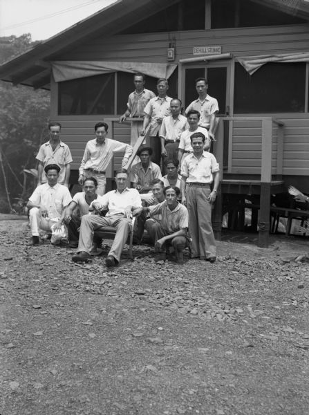 The camp construction executive and workers gather to pose for a group portrait in Panama. The executive is sitting in a chair and the workers are gathered around him sitting and standing on steps up to a cabin. The executive has a cigar in one hand and is wearing a light collar shirt and slacks. The workers are wearing similar clothing. Behind the cabin are woods. The group is multiracial.