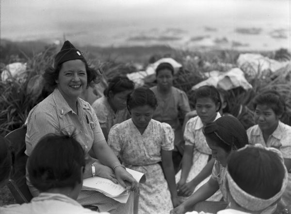 United States Army nurse, Mary Harrington, teaches female internees in Saipan, gathered outdoors in the grass with Saipan women. Harrington is smiling and holding a booklet. She is wearing a nurses' cap, matching striped shirt and pants, identification bracelet and a watch. The women are wearing patterned dresses and wear their hair up. There are plants and perhaps a shoreline in the background.