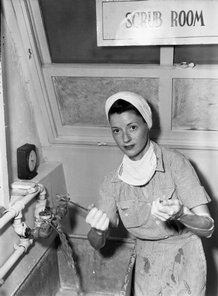 United States Army nurse, Ann T. Hyland, is washing her hands in the 148th General Hospital's scrub room. Hyland is wearing a white cloth around her hair, a striped pantsuit, and a surgeon's mask around her neck. She is holding up her arms, which are covered in soap. The water is running in the sink behind her, and the sign above her reads: "Scrub Room".