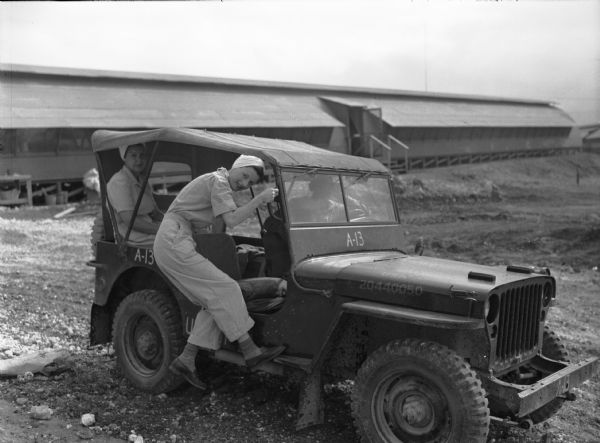 United States Army nurse, Ann T. Hyland, boarding a Jeep at 148th General Hospital in Saipan. Hyland is wearing a striped jumpsuit and has her hair tied up in a white cloth. There is a person in the Jeep's driver seat, and another female nurse is sitting in the back of the Jeep. In the background is a long building.