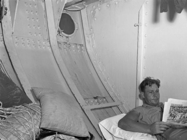 An off-duty member of the United Fruit Company's S.S. <i>Santa Marta</i> freighter reading a newspaper. The man is resting against the wall of the ship and is wearing work clothes. Beside him is a pillow and objects covered in a rope netting. This photograph was taken in the Caribbean.