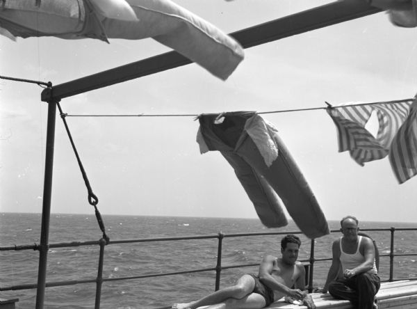 Two members of the United Fruit Company's S.S. <i>Santa Marta</i> sitting by the rail on a bench as laundry dries on a clothesline in the foreground. One man is lying down wearing shorts, and the man sitting next to him is wearing a sleeveless shirt, pants, and eyeglasses. This photograph was taken in the Caribbean.
