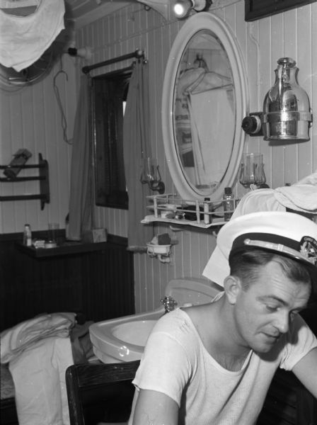 Off- duty shipmate on United Fruit Company's S.S. <i>Santa Marta</i>. The man is wearing a first mate hat and a white t-shirt. Behind him is a sink with a mirror above it and toiletries. Behind that is an unmade bed and a window. This photograph was taken in the Caribbean.