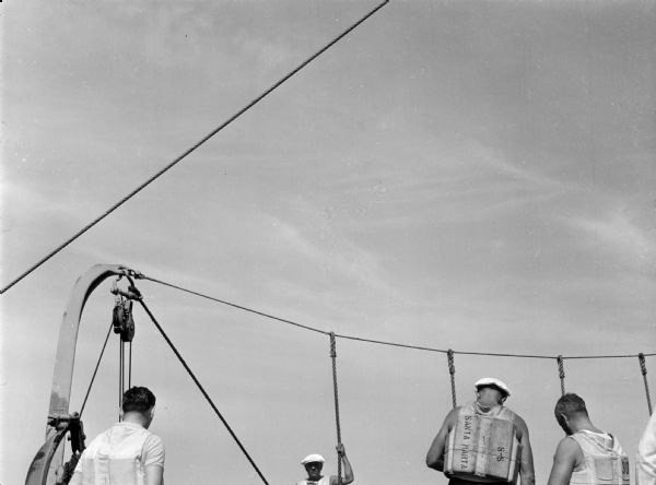 Sailors aboard the United Fruit Company's S.S. <i>Santa Marta</i> practice lifeboat drills in the Caribbean. All the men are wearing life jackets, and two of the men are wearing hats. Ropes from the freighter are visible against the sky.