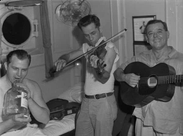 Men in a three piece orchestra, consisting of a guitar, a violin, and a glass jug, posing for a photograph aboard the United Fruit Company's S.S. <i>Santa Marta</I>. The three men are in the midst of playing their instruments. The man with the glass jug is not wearing a shirt and is holding a glass jug marked as sherry wine. Behind him on a bunk is the open violin case. The violin player has his bow raised and his violin under his chin, and is wearing a t-shirt and pants with a belt. The man playing guitar is wearing a loose button-up shirt and pants. This photograph was taken in the Caribbean.