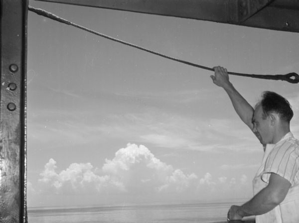 A crew member of the United Fruit Company's S.S. <i>Santa Marta</i> looking over the side of the freighter at the Caribbean Sea and the clouds. The man is holding onto a rope above his head with one hand. He is wearing a life jacket and a striped short-sleeved shirt.