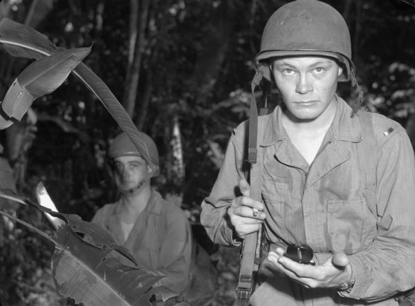 A patrol officer from the 14th Infantry, based in Fort Davis, Panama, follows his compass through the jungle. The patrol officer has a small cigarette in one hand and is holding his compass in front of him with his other hand. He is wearing fatigues and has his weapon's strap slung over his arm. Behind him is another soldier who is also wearing fatigues and carrying a machete.