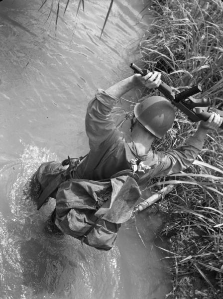 View from above of a soldier fording a stream with grassy banks near Fort Davis, Panama,  holding his rifle over his head. He is wearing fatigues and a backpack.