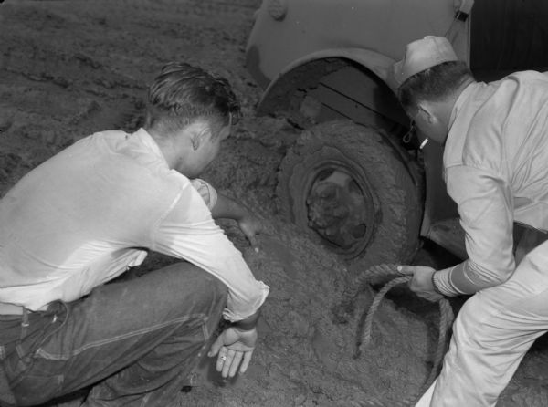 Soldiers from the 14th Infantry work to move a stuck Jeep from deep mud. One man in uniform with a cigarette in his mouth is leaning down over the wheel holding a rope. Another man in a shirt and pants is squatting down while pointing at the wheel. He is holding a cigarette in his hand and the back of his shirt is wet.
