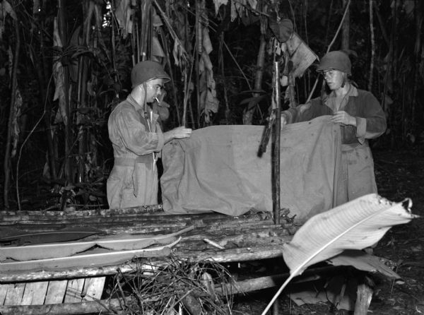 Two soldiers from the 14th Infantry building a "wiki-up", or place to sleep while on patrol, in a jungle in Panama. One man has a cigarette in his mouth.