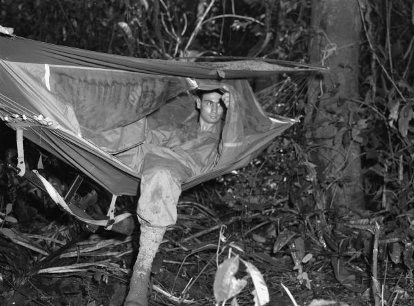 A soldier wearing fatigues in a "wiki-up", or place to sleep during patrol, in a jungle in Panama. The man is in a hammock which also has a mosquito net. His sub-machine gun is hanging below the hammock.