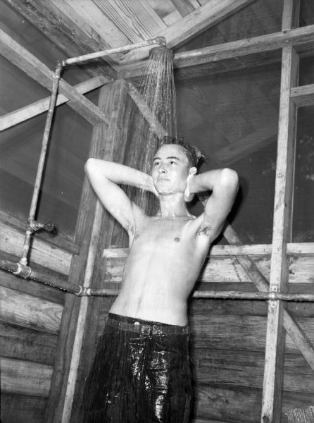 A soldier from the 14th Infantry taking a shower with his pants on at a military base in Panama. The soldier is shirtless and has his arms raised behind his head.