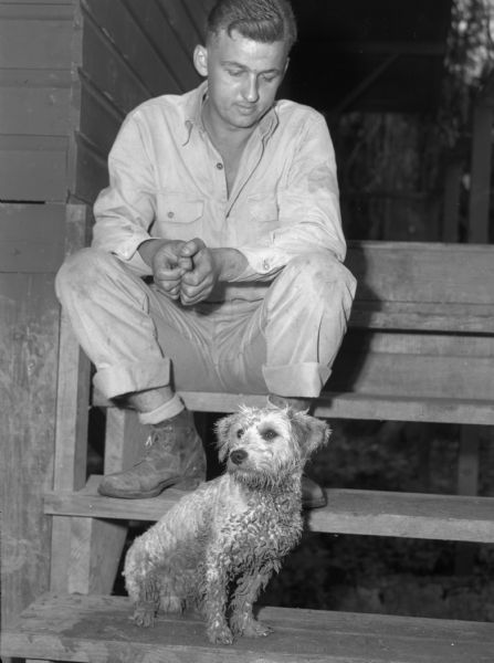 A soldier at a base in Panama sitting with the base's mascot, a small dog. The dog has matted, muddy fur and is sitting on wooden steps. A few steps above him is the soldier, wearing a button up shirt, rolled up pants, and boots. A cabin is behind him.