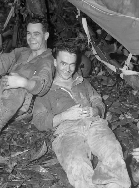 Two soldiers laughing and relaxing under a tree in Panama. One man is lying on his back and the other man is leaning against him. They are both wearing work uniforms. Beside one soldier is his sub-machine gun, hanging from a hammock, and a helmet behind him. Another sub-machine gun is propped against a tree by the other soldier.