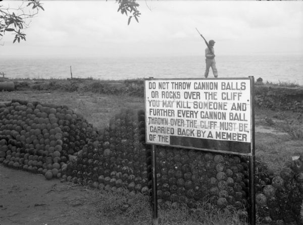 A soldier walks along the edge of a cliff in Panama, behind stacks of cannonballs and a large sign. The man is wearing a uniform and is holding a sub-machine gun. Beyond him is a cliff, with a large body of water in the background.