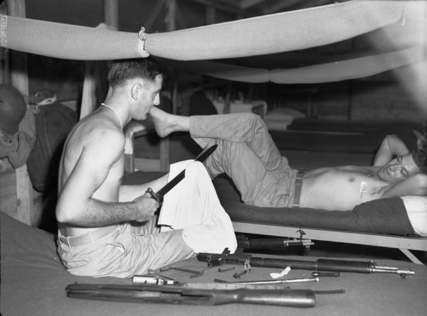 Two soldiers sitting and talking in Panama as one of them is cleaning his weapons. One man is sitting on a bed holding a machete and cleaning it with a towel. Next to him is a disassembled rifle. He is shirtless and looking at man who is lying on a bed. Above them are hanging rolled-up mosquito nets.