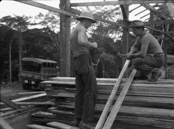 Two soldiers from the 14th Infantry working among lumber in Panama. One man iswearing a hat and squatting on top of a pile of lumber while holding a hammer. Another shirtless man wearing a hat is standing atop another pile of lumber. He is holding a piece of wood and a tool. Above them is the frame of a building.