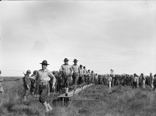 A group of men from the 14th Infantry running while crossing a newly constructed wooden bridge over mud in a field in Panama. The men are in uniform and are being watched by a group of men, also in uniform. There are mountains in the far background.