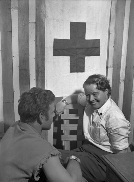 Martha Pratt, assistant field director at the Red Cross hospital in Saipan, participates in conversational therapy with a soldier wounded in Iwo Jima. Pratt is sitting facing the soldier, and is wearing a white button-up shirt with a cross on the collar. Behind her is a Red Cross banner. The soldier is facing her and wearing a shirt with torn off sleeves.