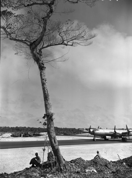 A B-29 bomber plane landing at North Field in Guam after its first fire raid on Tokyo. A tree is in the center foreground with three men sitting around it watching the plane land. On the other side of the air strip are low buildings with pointed roofs and two trucks.