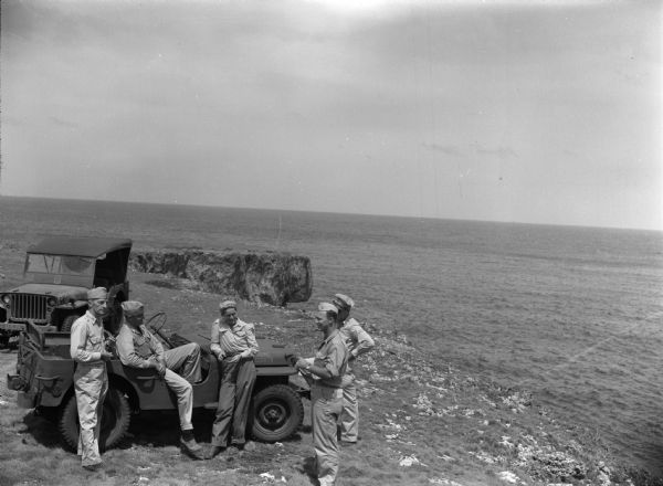 Leota Kelly, directer of the Pacific Athletic Club, division of the Red Cross, and colleagues, at the site of the new Red Cross Club in Tinian near a cliff with the ocean in the background. One man is leaning against a Jeep in which one man is sitting in the passenger seat. Kelly is standing next to him and two other men are standing off to the right side. She is wearing pants and a long-sleeved, button-up shirt with her hair tied up in a cloth. There is another Jeep in the background.